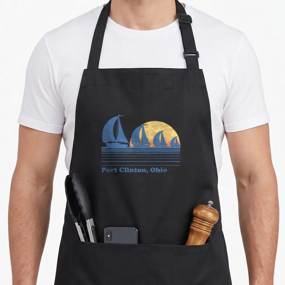 Port 80s apron - Four blue sailboats before yellow sun with Port Clinton Ohio text and horizontal blue water lines — shown on a black cotton canvas apron against a white background