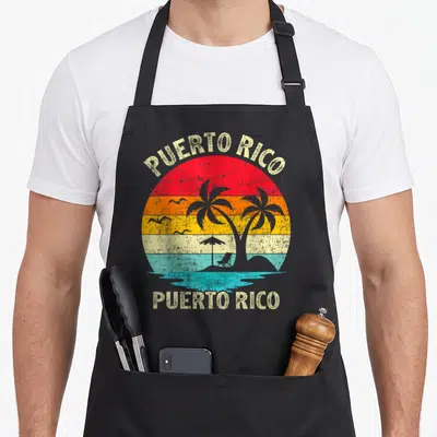 Puerto family aprons - Retro circular sunset with palm trees, beach chair, umbrella, birds, and text PUERTO RICO above — shown on a black cotton canvas apron against a white background