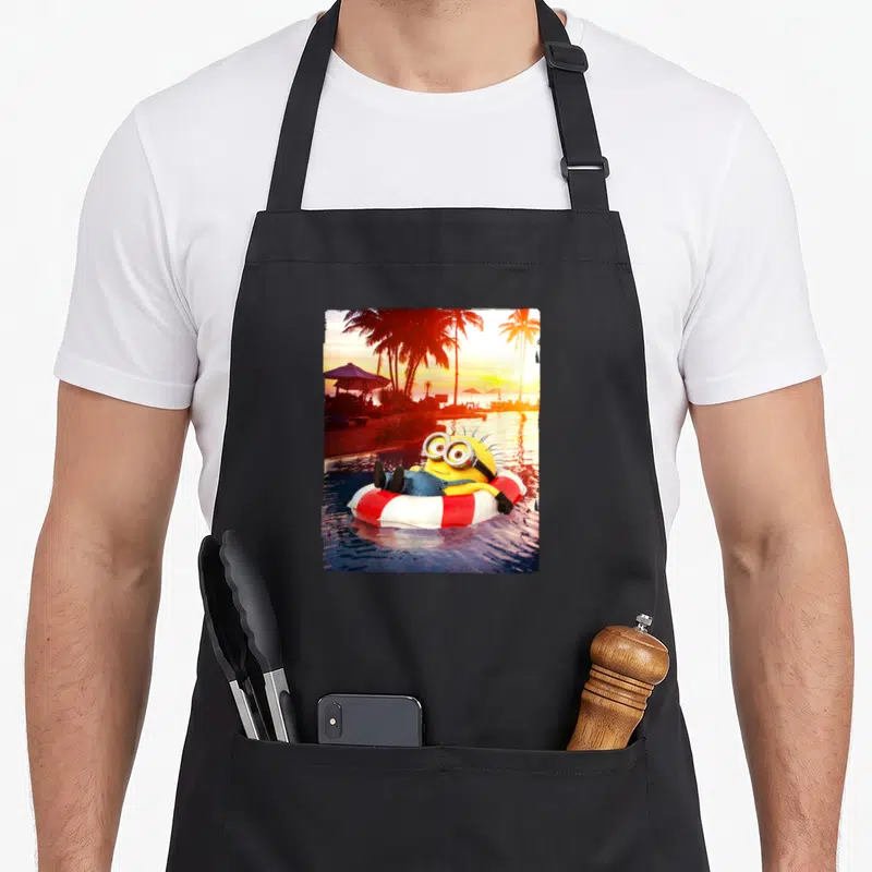 Jerry despicable me apron - Yellow minion in red white float ring tropical sunset palm trees — shown on a black cotton canvas apron against a white background