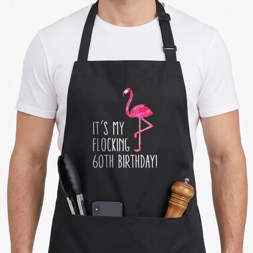 60th pink flamingo apron - Watercolor pink flamingo standing beside white text IT'S MY FLOCKING 60TH BIRTHDAY! — shown on a black cotton canvas apron against a white background