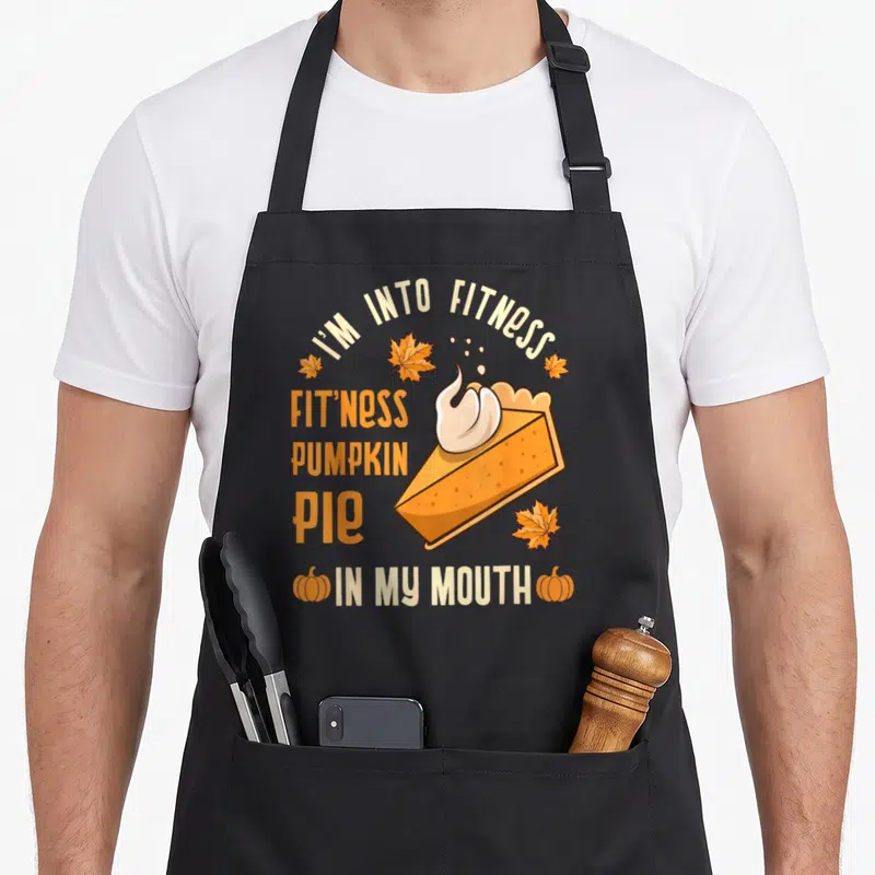 Cool turkey thanksgiving day aprons - Cartoon pumpkin pie slice with whipped cream and fitness pun text surrounded — shown on a black cotton canvas apron against a white background