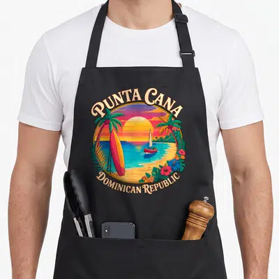 Republic vacation punta cana apron - Retro circular Punta Cana sunset with sailboat, surfboard, and hibiscus flowers — shown on a black cotton canvas apron against a white background