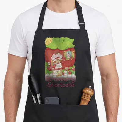 Poster strawberry apron - Cartoon girl in doorway of red berry house with cat in window and centered text below white — shown on a black cotton canvas apron against a white background