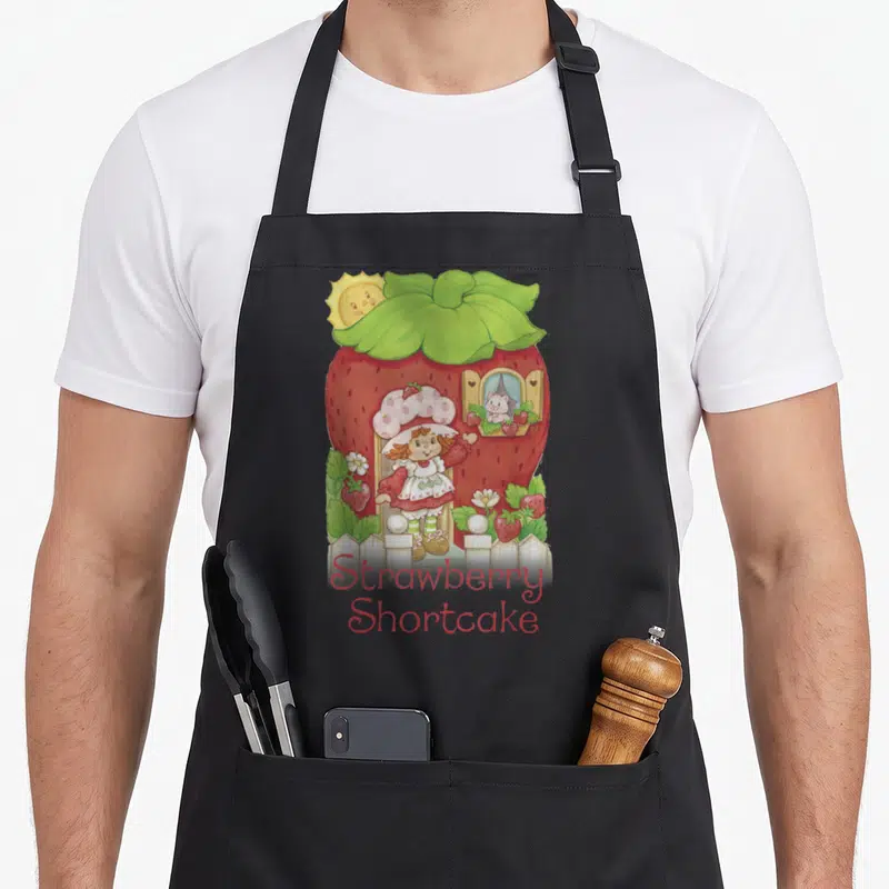 Poster strawberry apron - Cartoon girl in doorway of red berry house with cat in window and centered text below white — shown on a black cotton canvas apron against a white background