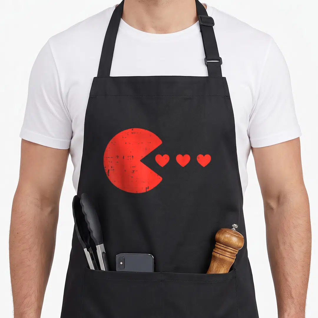 Eating valentine's day apron - Red Pac-Man figure facing three small red hearts in a row — shown on a black cotton canvas apron against a white background