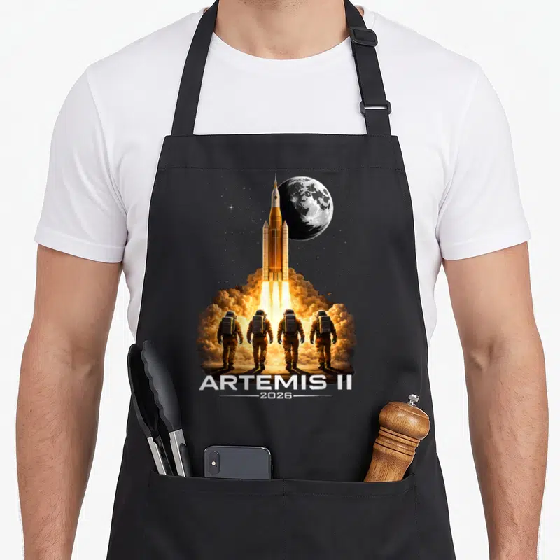 Artemis boy astronaut apron - Orange rocket launching vertically with four astronauts and ARTEMIS II 2026 text — shown on a black cotton canvas apron against a white background