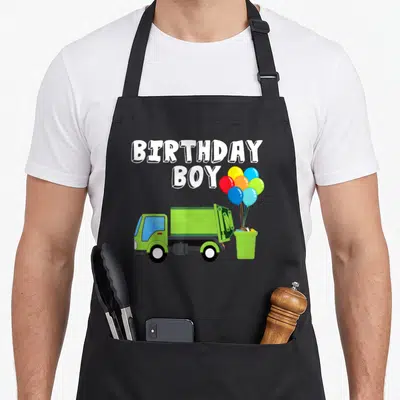 Balloon truck apron - Green garbage truck and colorful balloons with BIRTHDAY BOY text in cartoon style — shown on a black cotton canvas apron against a white background