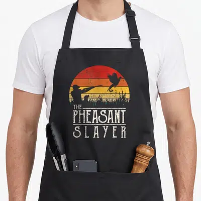 Sunset slayer apron - Retro striped sunset with hunter silhouette and THE PHEASANT SLAYER text — shown on a black cotton canvas apron against a white background