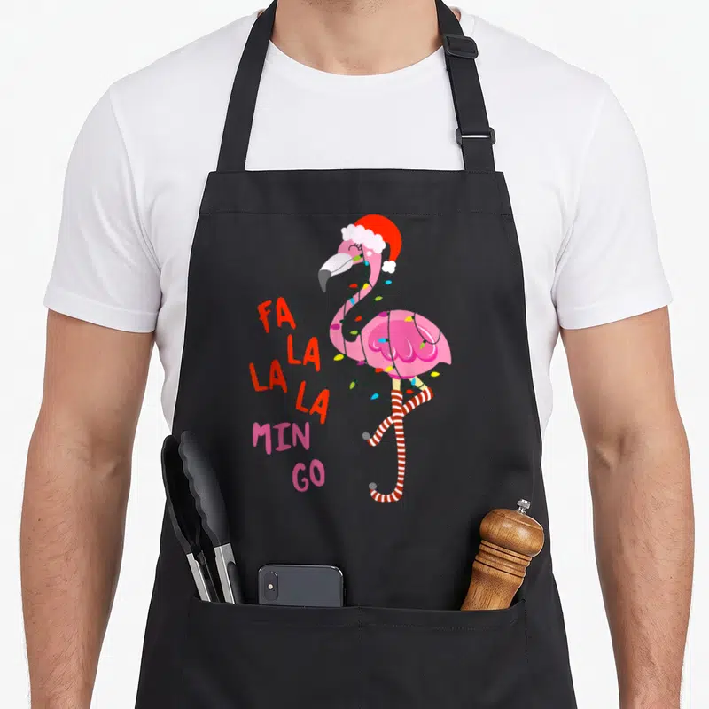 Mingo flamingo apron - Pink flamingo wearing red santa hat and lights with stacked fa la la min go text — shown on a black cotton canvas apron against a white background