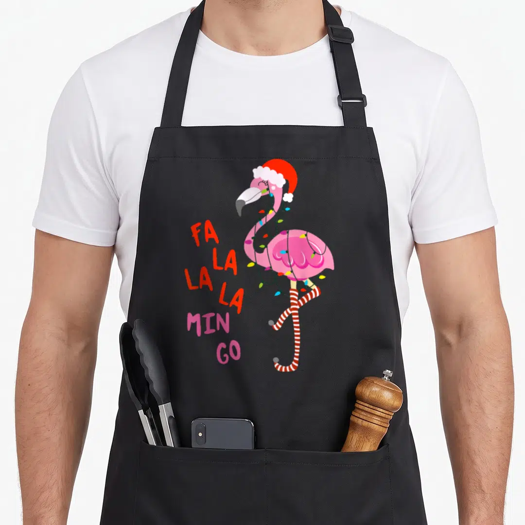 Mingo flamingo apron - Pink flamingo wearing red santa hat and lights with stacked fa la la min go text — shown on a black cotton canvas apron against a white background