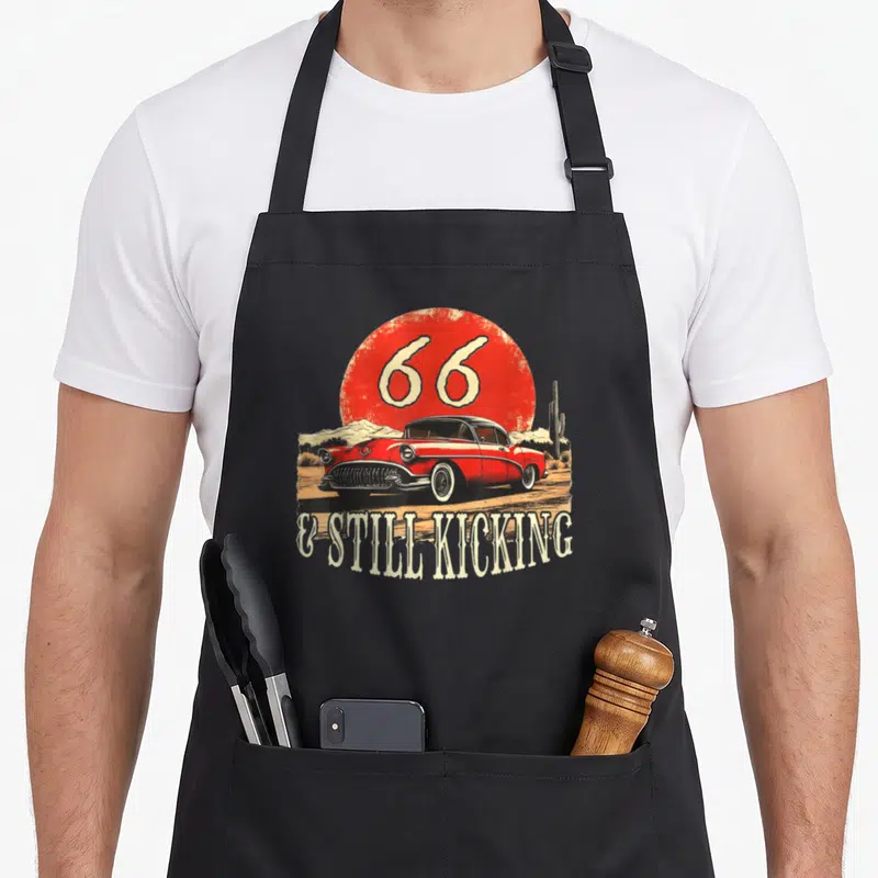 Route mother's apron - Red vintage car under 66 sunburst with text & still kicking — shown on a black cotton canvas apron against a white background