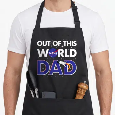 Father nasa apron - Out of This World NASA Dad text with blue planet logo and flying space shuttle — shown on a black cotton canvas apron against a white background