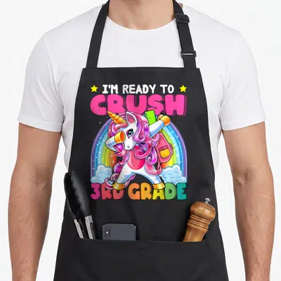 3rd unicorn apron - Dabbing unicorn holding ruler and pencil under rainbow with I'm ready to crush 3rd grade text — shown on a black cotton canvas apron against a white background