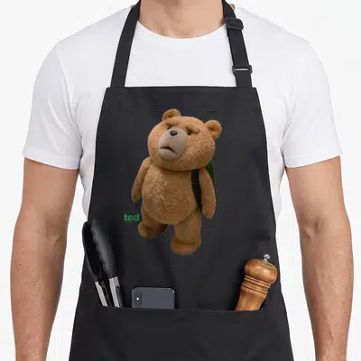 Ted teddy bear apron - Brown teddy bear wearing dark green backpack with green ted logo text — shown on a black cotton canvas apron against a white background
