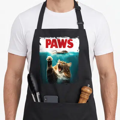 Humorous cat apron - Red PAWS text above swimming mouse and reaching cat in blue water — shown on a black cotton canvas apron against a white background