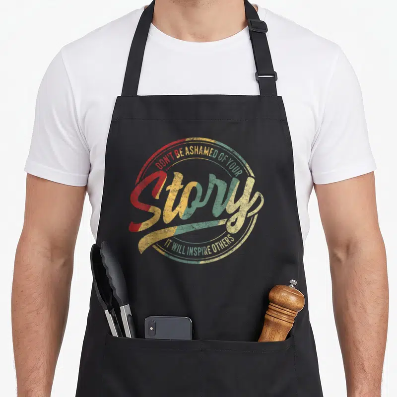 Recovery story of the apron - Retro circular text design reading Don't Be Ashamed Of Your Story It Will Inspire Others — shown on a black cotton canvas apron against a white background
