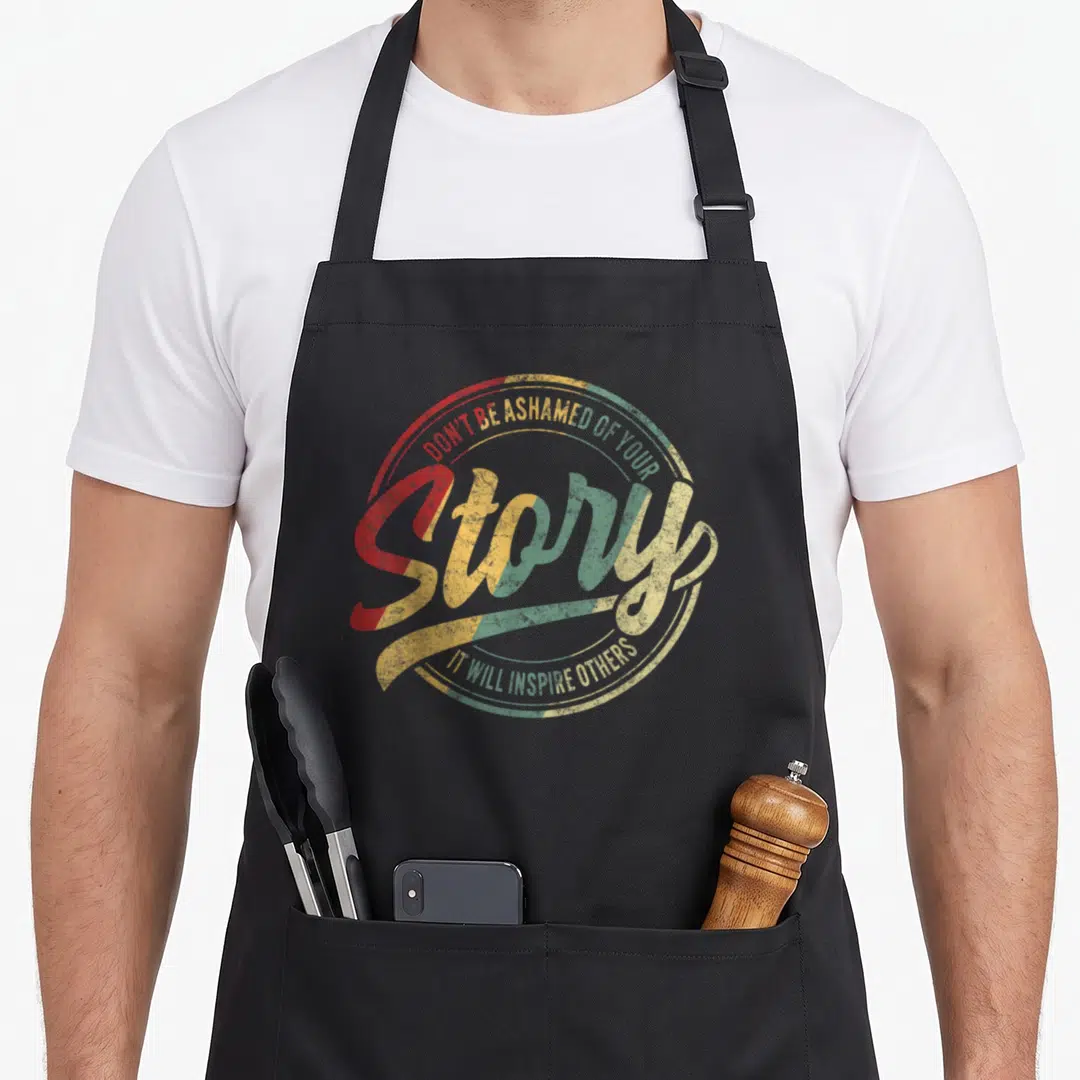 Recovery story of the apron - Retro circular text design reading Don't Be Ashamed Of Your Story It Will Inspire Others — shown on a black cotton canvas apron against a white background