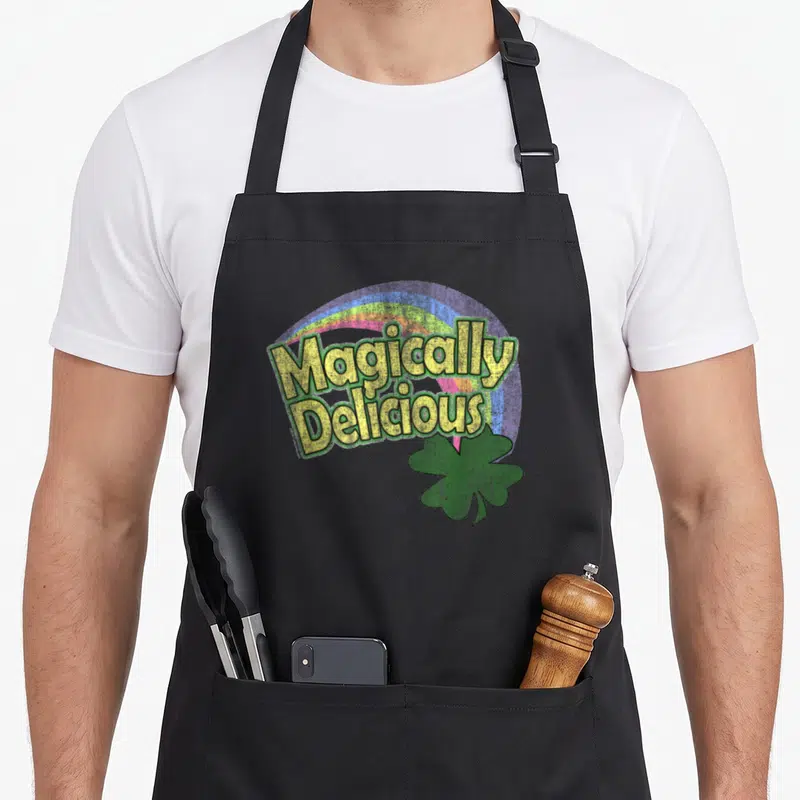 Cutecomfy st patrick day aprons - Stacked yellow text Magically Delicious over rainbow arch with green shamrock bottom — shown on a black cotton canvas apron against a white background