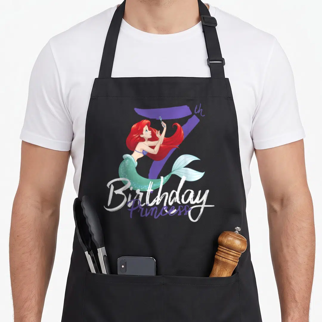 Ariel 7th princess apron - Red-haired mermaid holding fork in front of purple number 7 with Birthday Princess text — shown on a black cotton canvas apron against a white background