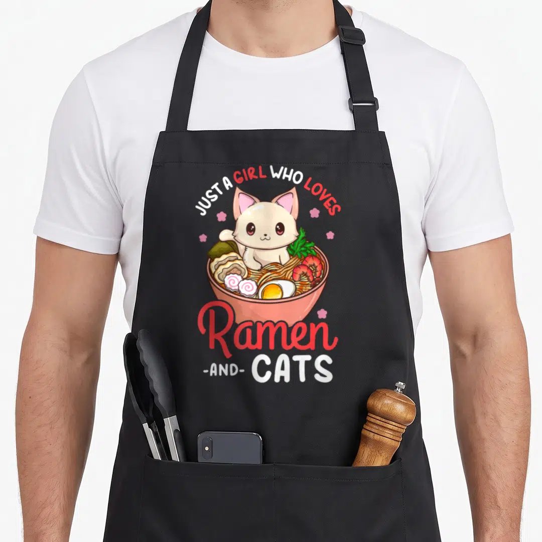 Neko anime cat apron - Cartoon cream cat in pink ramen bowl with text JUST A GIRL WHO LOVES Ramen AND CATS — shown on a black cotton canvas apron against a white background