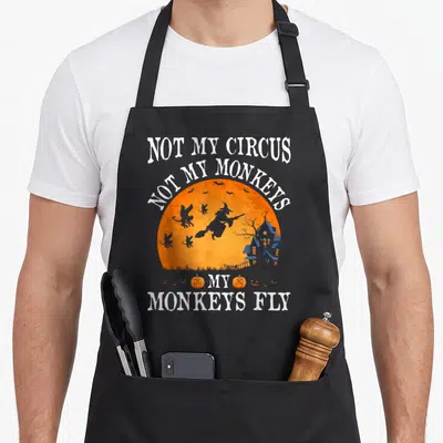 Circus flying apron - Witch flying past bats inside orange moon with Not My Circus text and pumpkins — shown on a black cotton canvas apron against a white background
