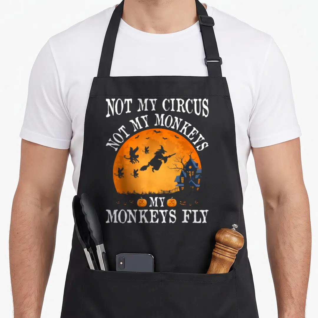 Circus flying apron - Witch flying past bats inside orange moon with Not My Circus text and pumpkins — shown on a black cotton canvas apron against a white background