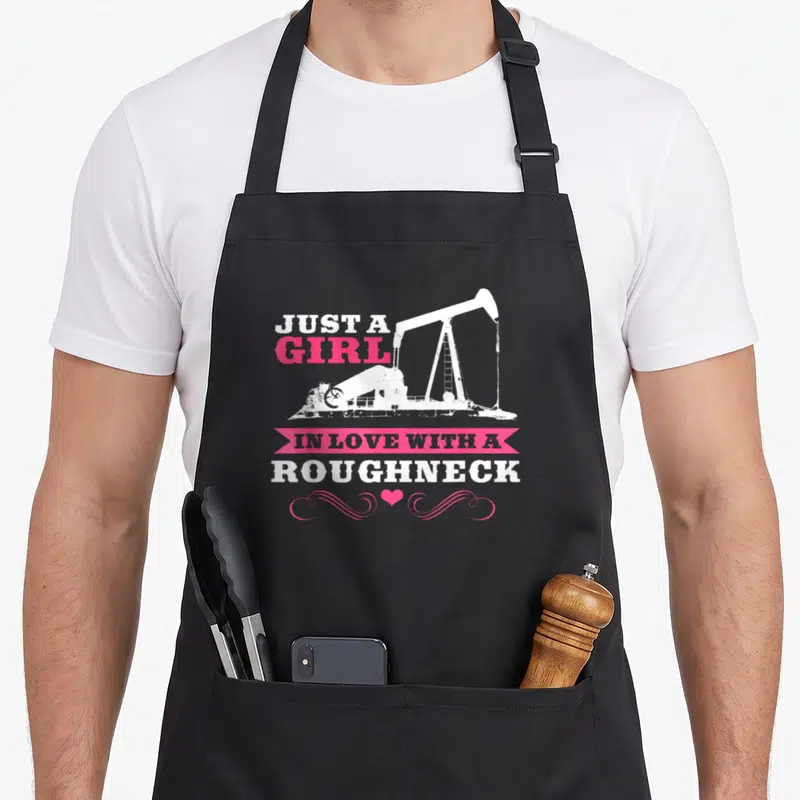 Rig wife apron - White pumpjack graphic with pink ribbon banner and distressed text reading JUST A GIRL IN LOVE — shown on a black cotton canvas apron against a white background