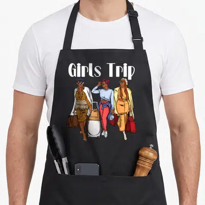 Melanin girls trip apron - Three women standing with luggage and shopping bags under white Girls Trip text — shown on a black cotton canvas apron against a white background