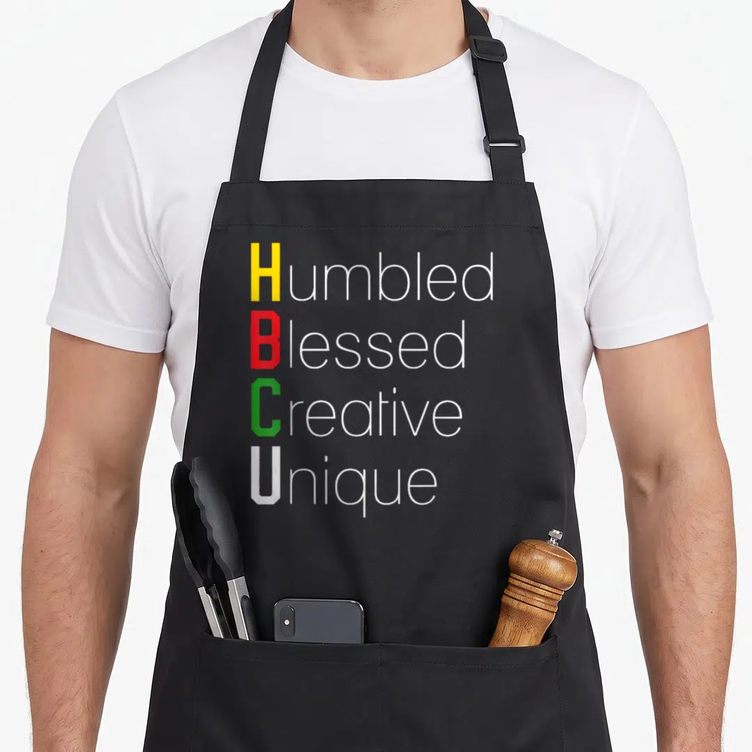 Hbcu creative apron - Stacked text Humbled Blessed Creative Unique with yellow red green and white initials — shown on a black cotton canvas apron against a white background