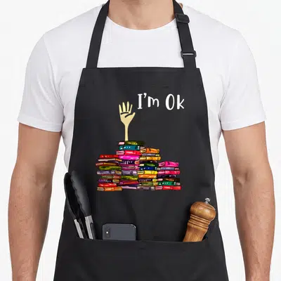 Reader teacher apron - I'm Ok text above hand reaching from multicolored book stack — shown on a black cotton canvas apron against a white background
