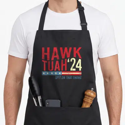 Hawk viral apron - Stacked red and white text reading HAWK TUAH 24 above blue flag banner and blue phrase Spit On That — shown on a black cotton canvas apron against a white background