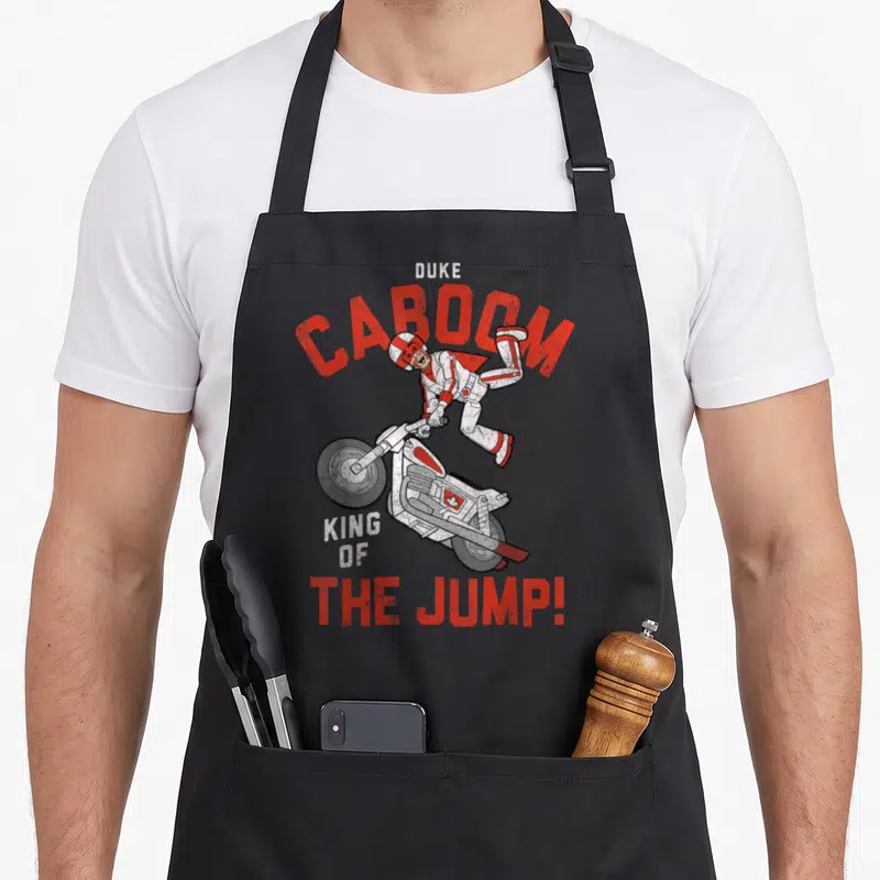 Duke king apron - Man jumping above motorcycle with Duke Caboom King of the Jump text in red white grey — shown on a black cotton canvas apron against a white background