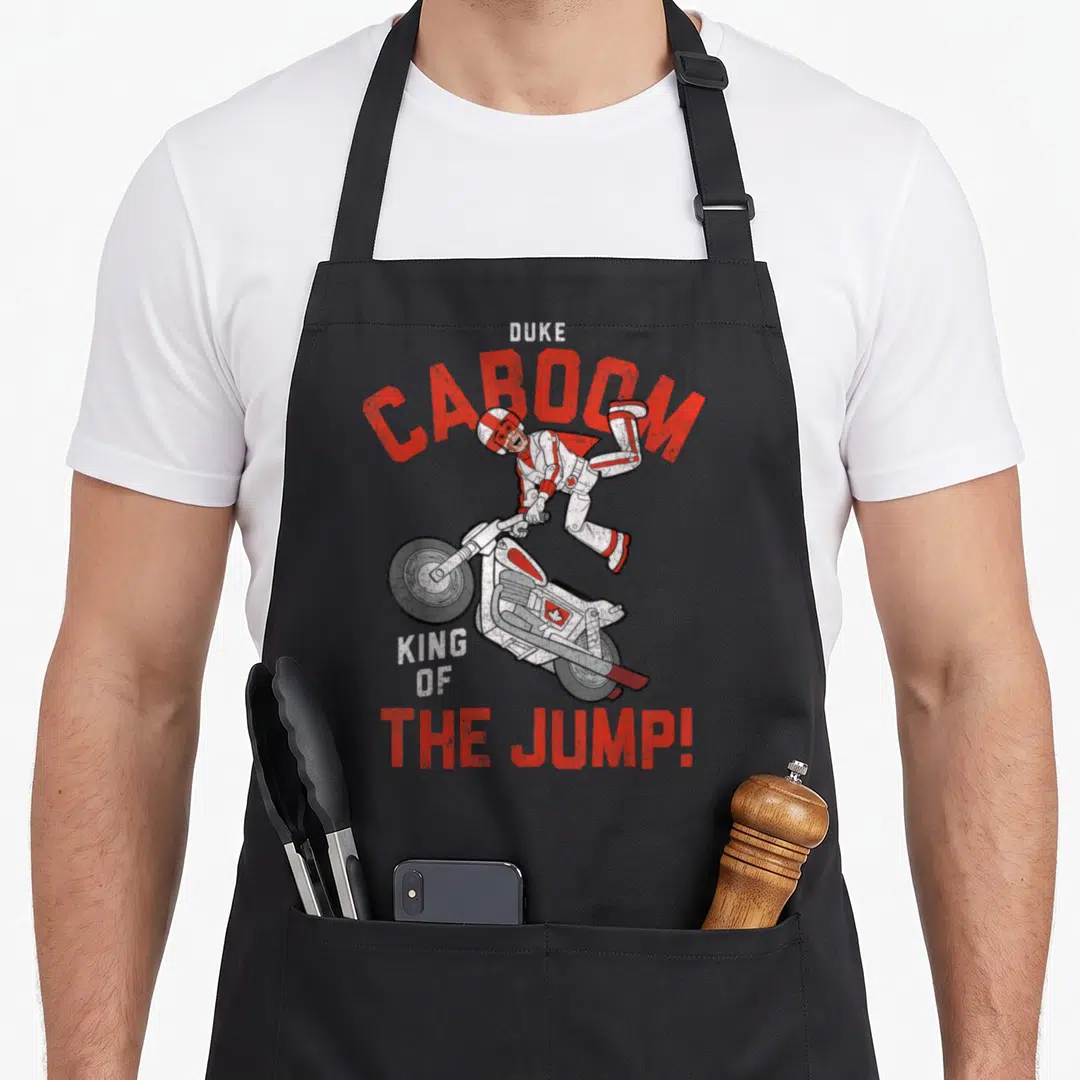 Duke king apron - Man jumping above motorcycle with Duke Caboom King of the Jump text in red white grey — shown on a black cotton canvas apron against a white background