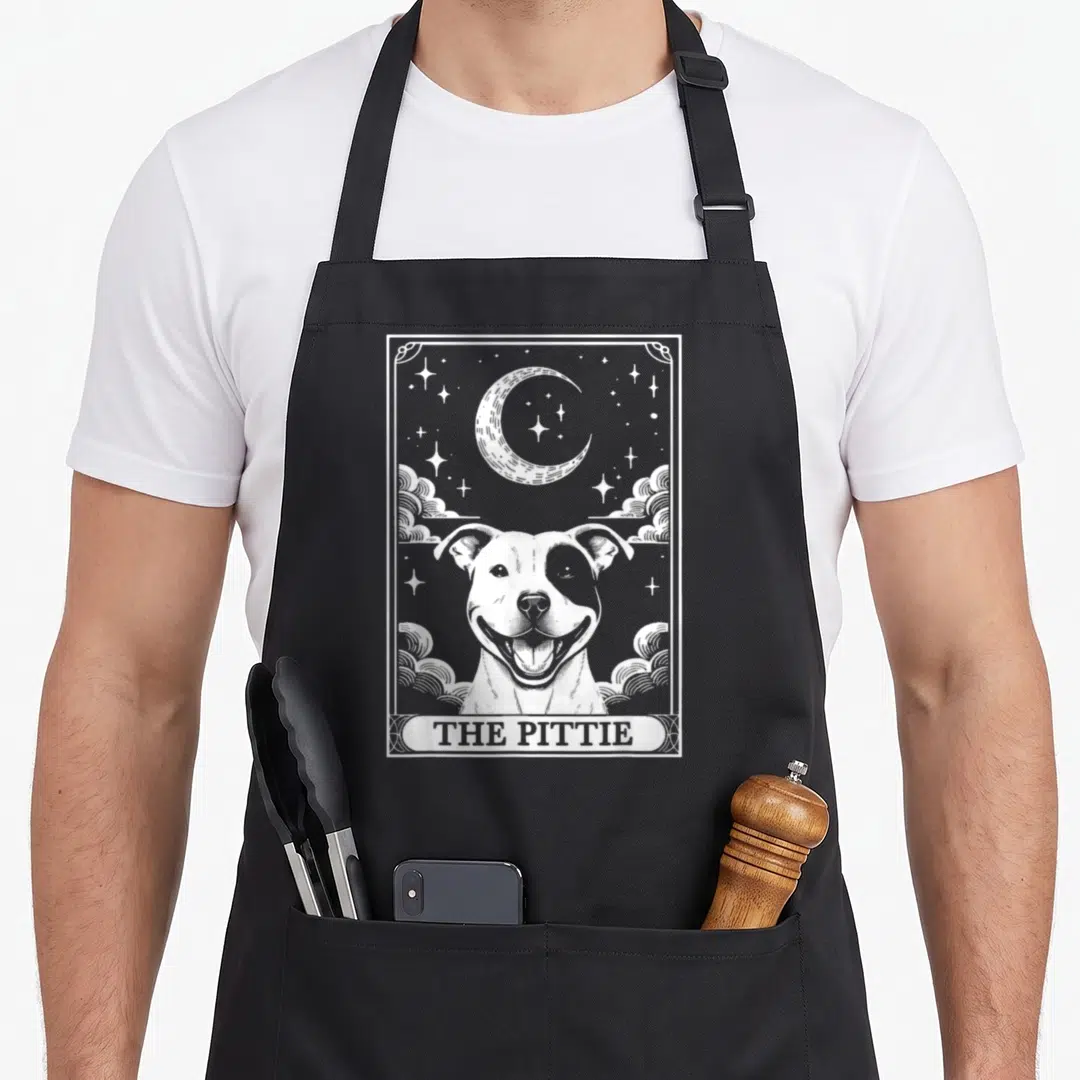 Tarot dog apron - White line art of smiling dog under crescent moon and stars with THE PITTIE text in decorative border — shown on a black cotton canvas apron against a white background