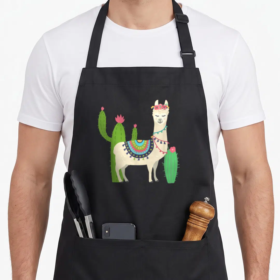 Llama cactus apron - Cartoon cream llama with floral crown and patterned blanket surrounded by three green cacti — shown on a black cotton canvas apron against a white background