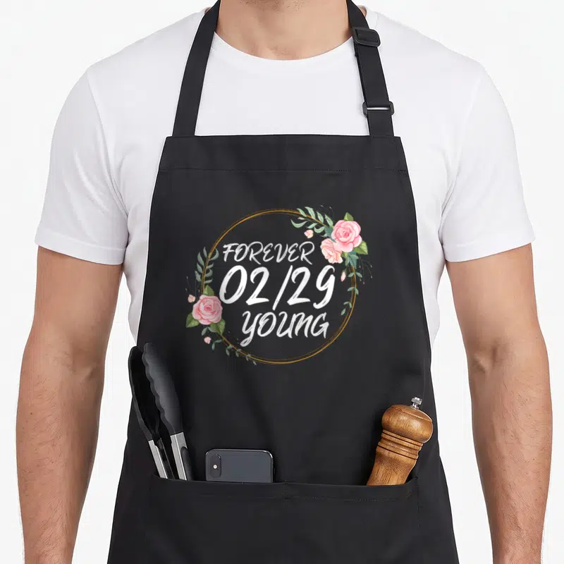Leap garland apron - White text forever 02/29 young in gold circle surrounded by pink roses and green leaves — shown on a black cotton canvas apron against a white background