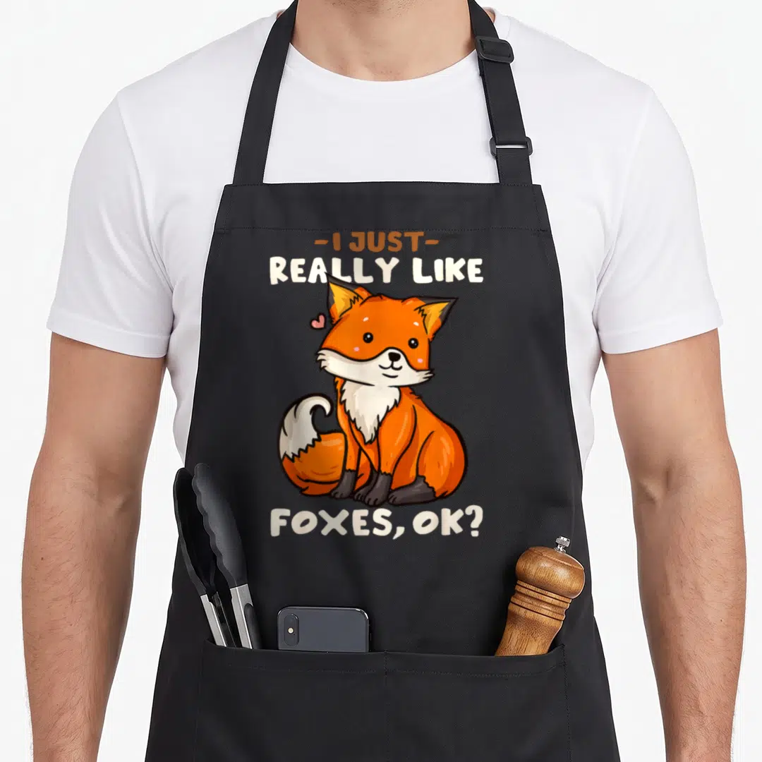 Cute fox aprons - Cartoon orange fox sitting with pink heart and text I Just Really Like Foxes Ok — shown on a black cotton canvas apron against a white background