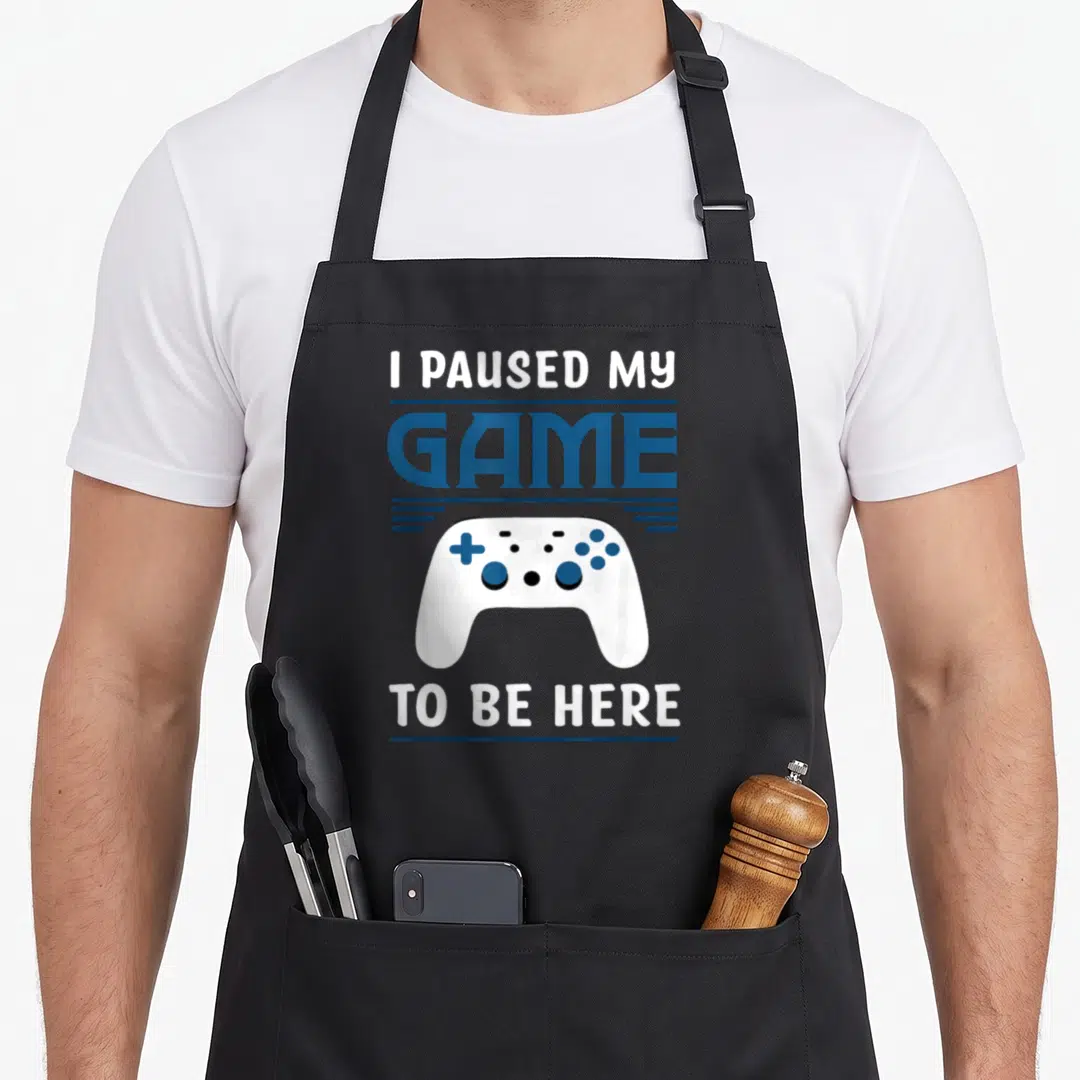 Paused video game apron - White game controller between blue and white text reading I Paused My Game To Be Here — shown on a black cotton canvas apron against a white background