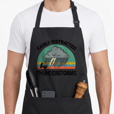Slightly thunderstorms apron - Retro striped circle with grey rain cloud, yellow lightning, and black text easily — shown on a black cotton canvas apron against a white background