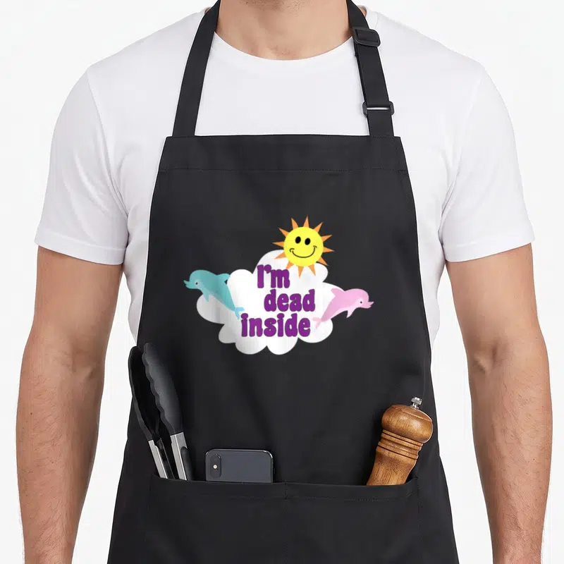 Dolphin goth apron - Purple I'm dead inside text in white cloud with blue and pink dolphins and yellow smiling sun — shown on a black cotton canvas apron against a white background