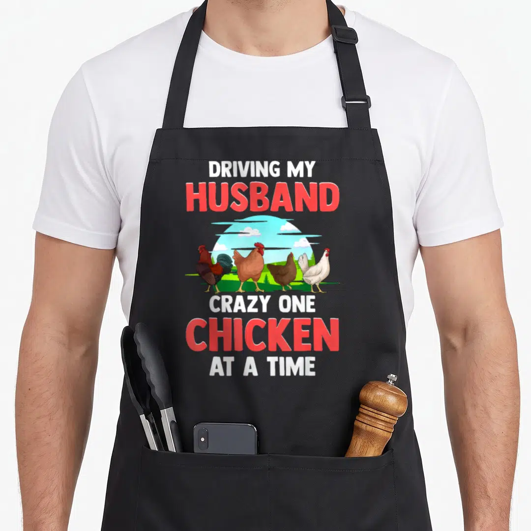 Chicken apron gift - Four cartoon chickens under blue sun with red and white text about driving husband crazy — shown on a black cotton canvas apron against a white background