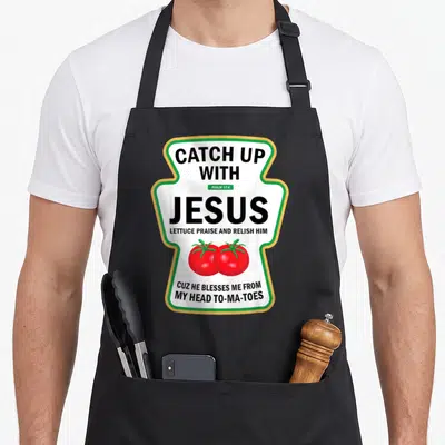 Pun catch up with jesus apron - Ketchup bottle shape with black text and two red tomatoes inside green and yellow border — shown on a black cotton canvas apron against a white background