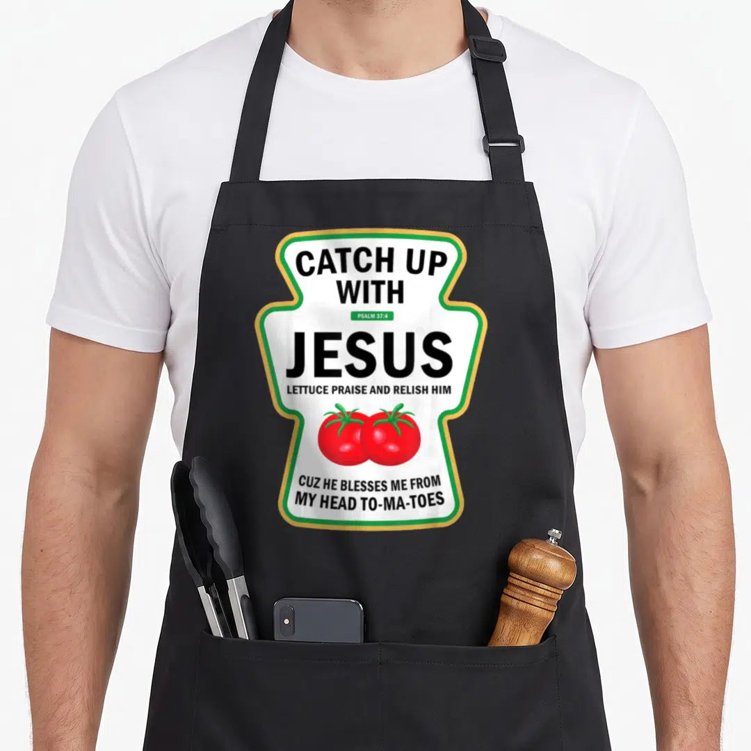Pun catch up with jesus apron - Ketchup bottle shape with black text and two red tomatoes inside green and yellow border — shown on a black cotton canvas apron against a white background