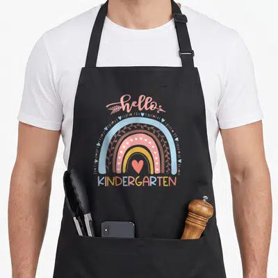 School hello kindergarten teacher apron - Cartoon rainbow with pink and blue bands, hearts, and hello kindergarten text — shown on a black cotton canvas apron against a white background
