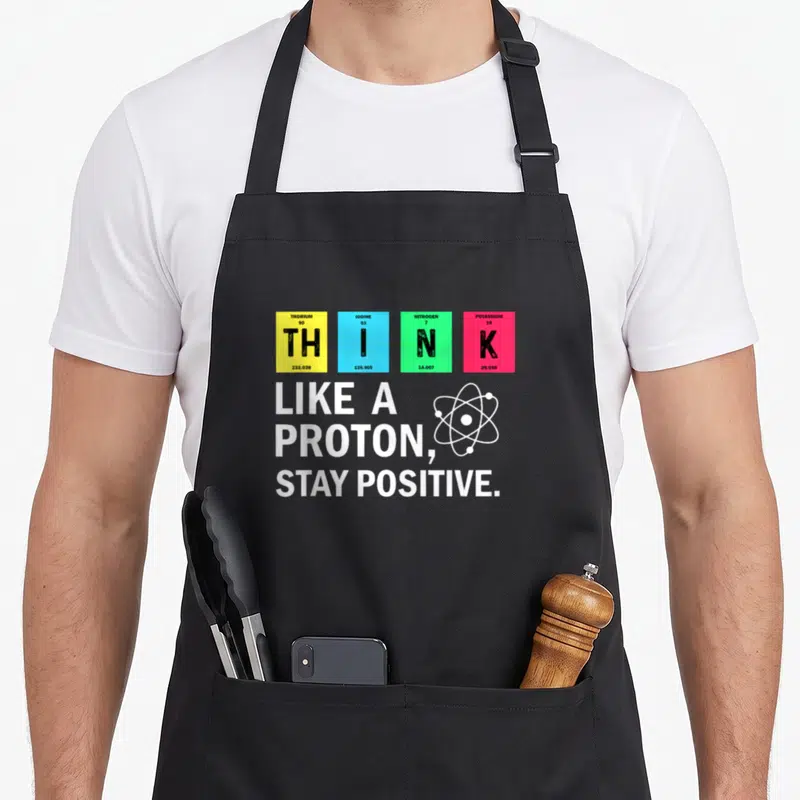 Proton science apron - Periodic table tiles spelling THINK above white text LIKE A PROTON STAY POSITIVE and atom symbol — shown on a black cotton canvas apron against a white background