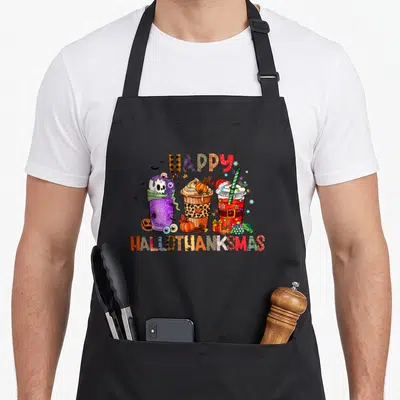Latte thanksgiving apron - Three holiday coffee cups with skull pumpkin and santa garnishes under happy hallothanksmas — shown on a black cotton canvas apron against a white background