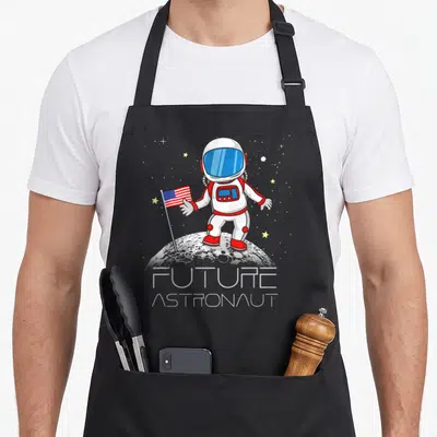 Future boy astronaut apron - Cartoon astronaut holding a flag on the moon with FUTURE ASTRONAUT text below — shown on a black cotton canvas apron against a white background
