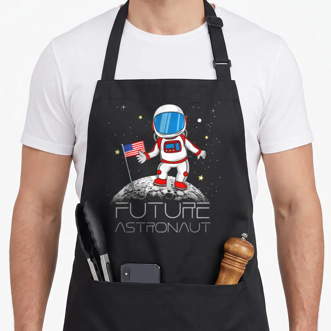 Future boy astronaut apron - Cartoon astronaut holding a flag on the moon with FUTURE ASTRONAUT text below — shown on a black cotton canvas apron against a white background