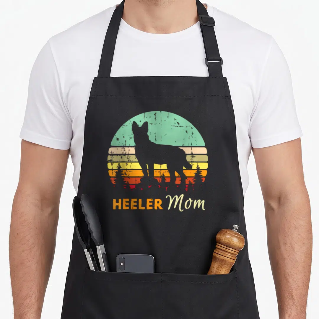 Heeler mother's apron - Grey dog silhouette standing before a retro striped sunset with HEELER Mom text below — shown on a black cotton canvas apron against a white background