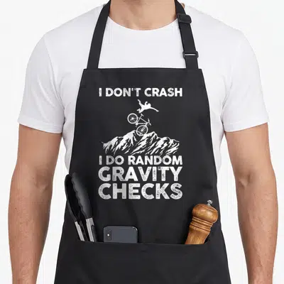 Mtb biking bike apron - White distressed text I Don't Crash Random Gravity Checks with airborne biker over mountain peak — shown on a black cotton canvas apron against a white background